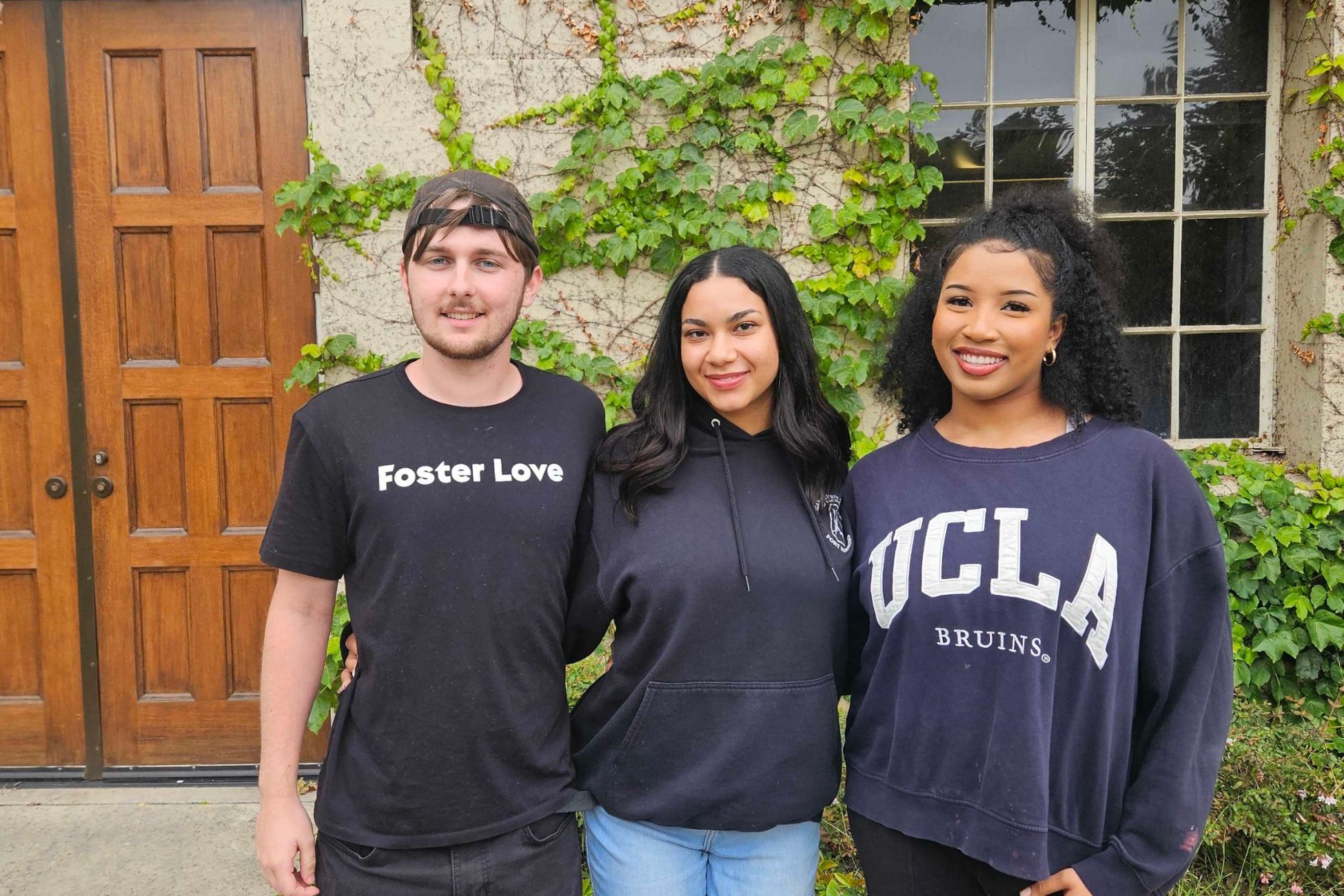 Three individuals stand in front of a building, smiling and wearing attire with &quot;UCLA&quot; and &quot;foster love&quot;.