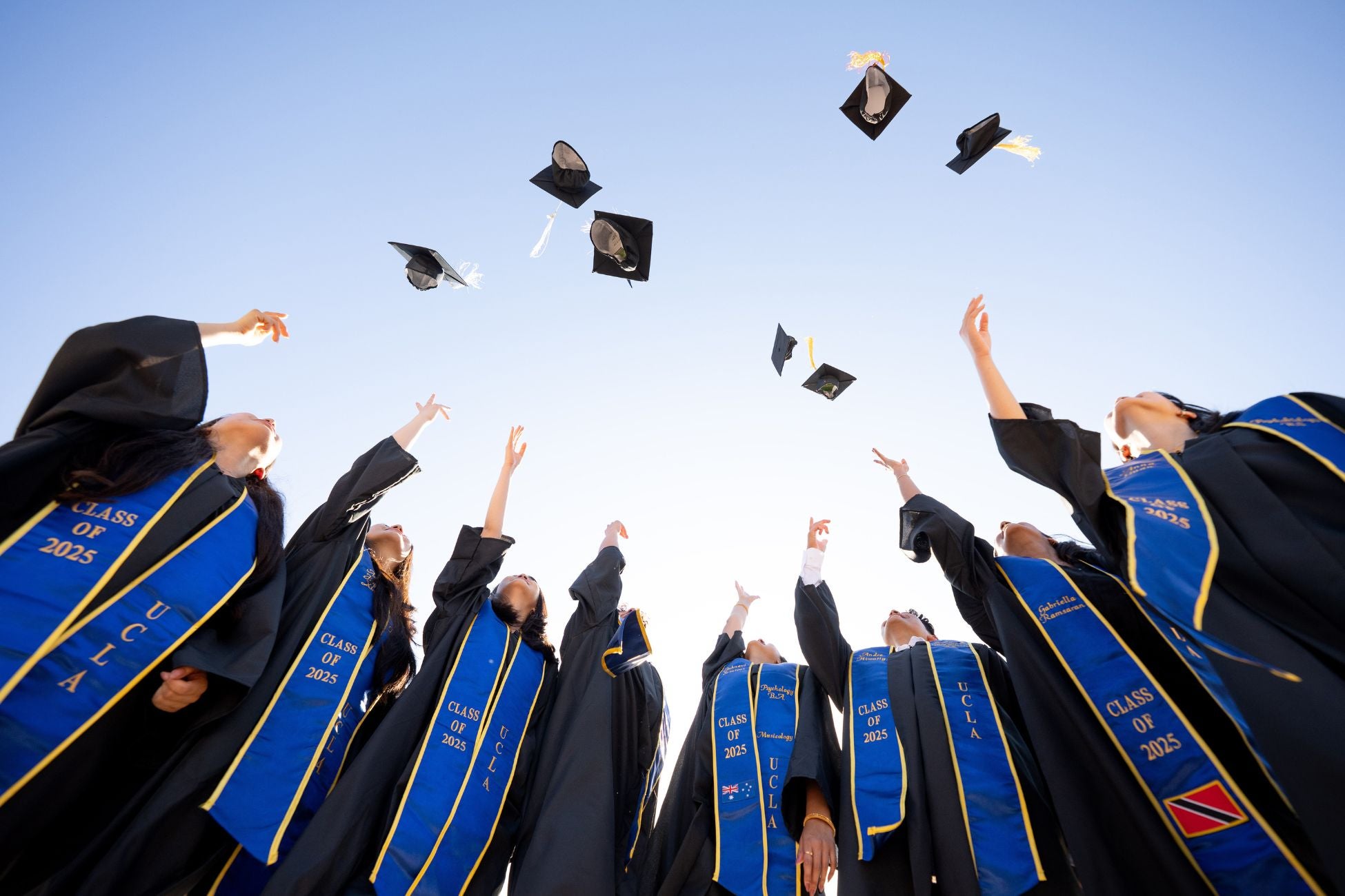 Group of students wearing graduation gowns and sashes stand in a circle throwing their caps in the air