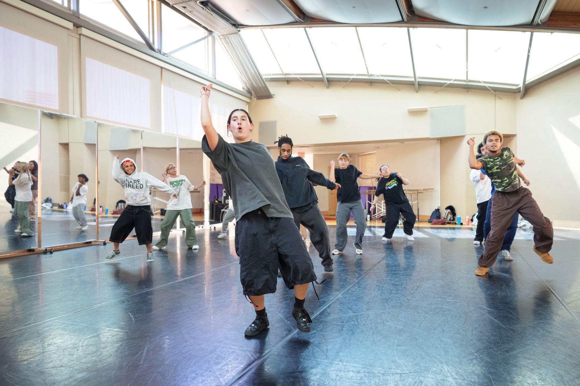 A group of dancers of various genders practice hip-hop in three rows next to a mirrored wall 