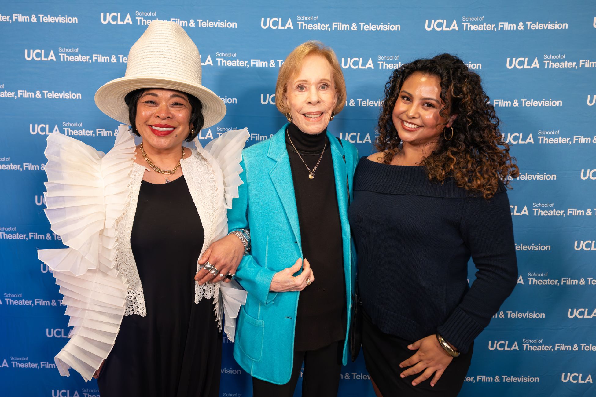 Three women pose and smile in front of a UCLA School of Theater, Film &amp; Television backdrop. 