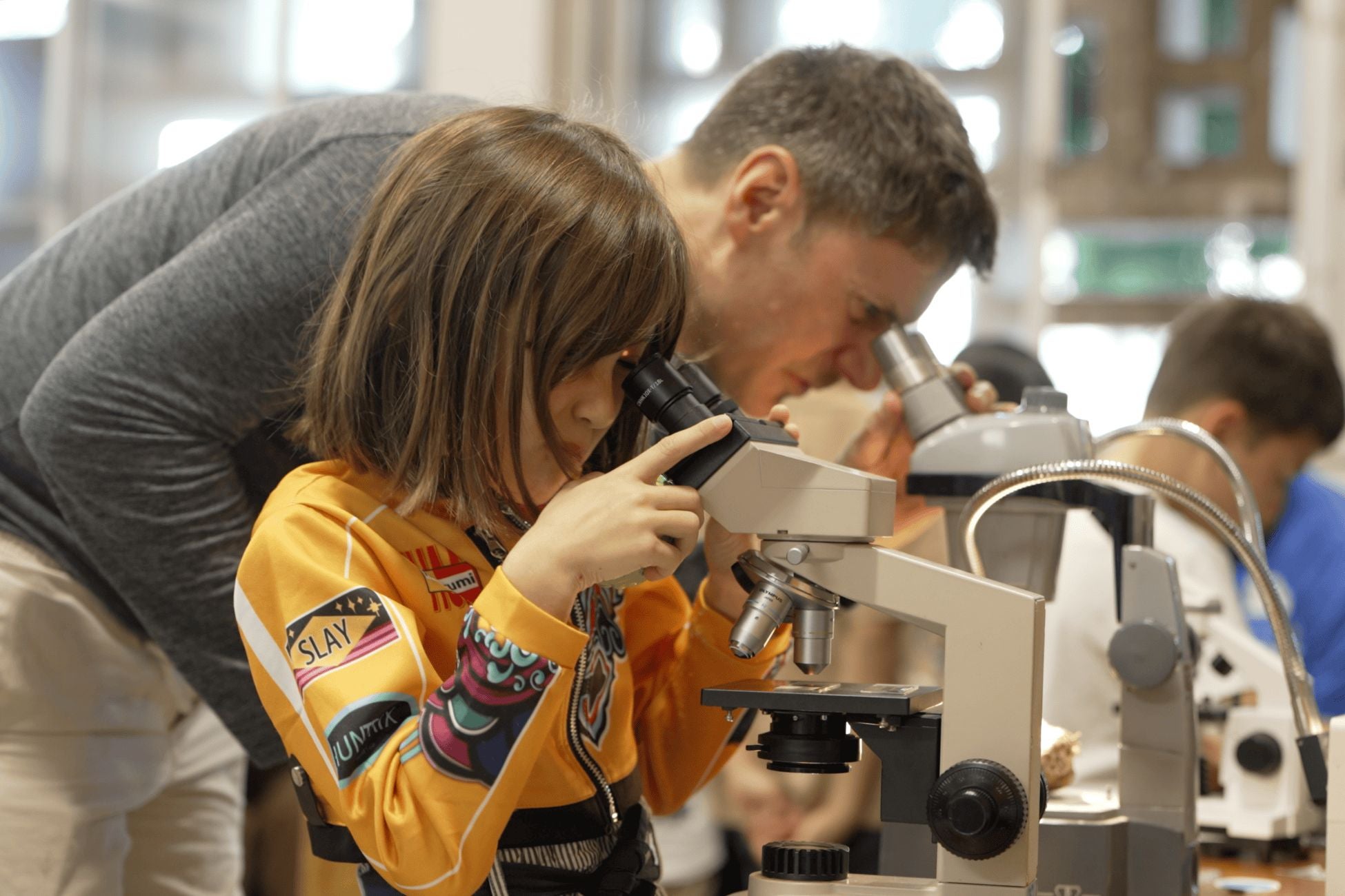 A man and a young girl observe specimens through microscopes, engaged in a scientific exploration together.