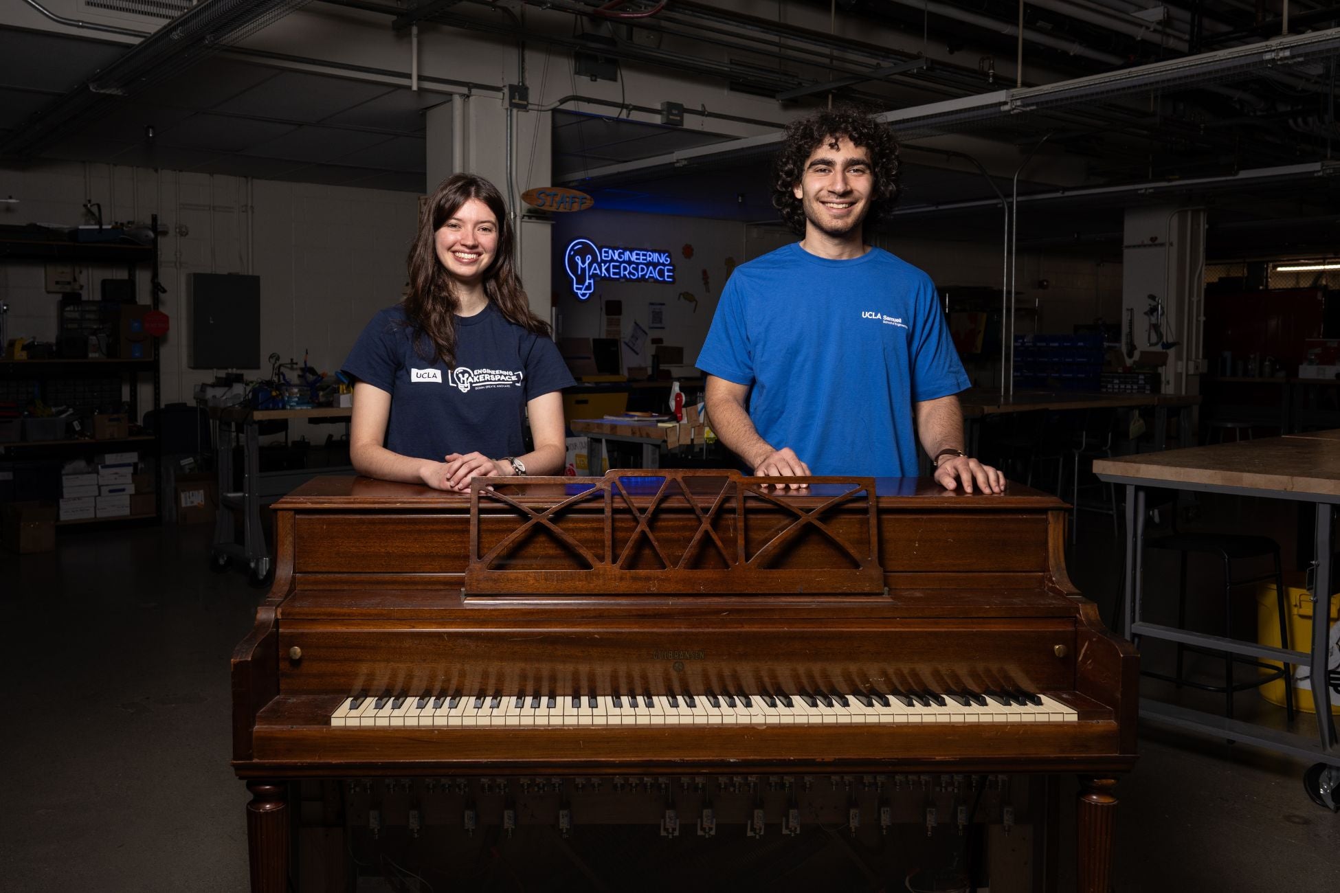 A young woman and young man stand smiling behind a brown piano. She has long brown hair and a navy blue shirt. He has short curly brown hair and a bright blue shirt. A neon sign glows behind them that reads "Engineering Makerspace".