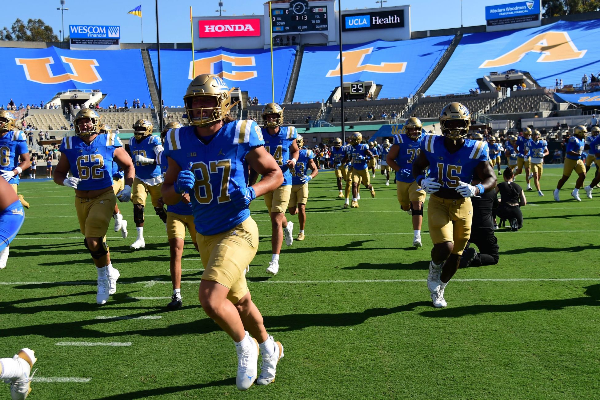 UCLA football players in blue and gold uniforms running onto the field before a game.