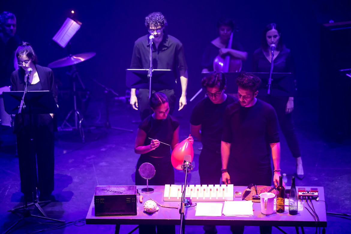 Performers on stage during a theatrical performance, with actors gathered around a table under purple lighting.
