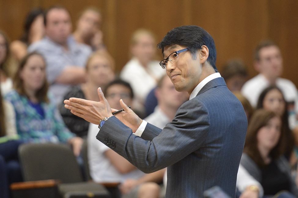 Korean American man with glasses and grey pinstripe suit gestures before group seated in background