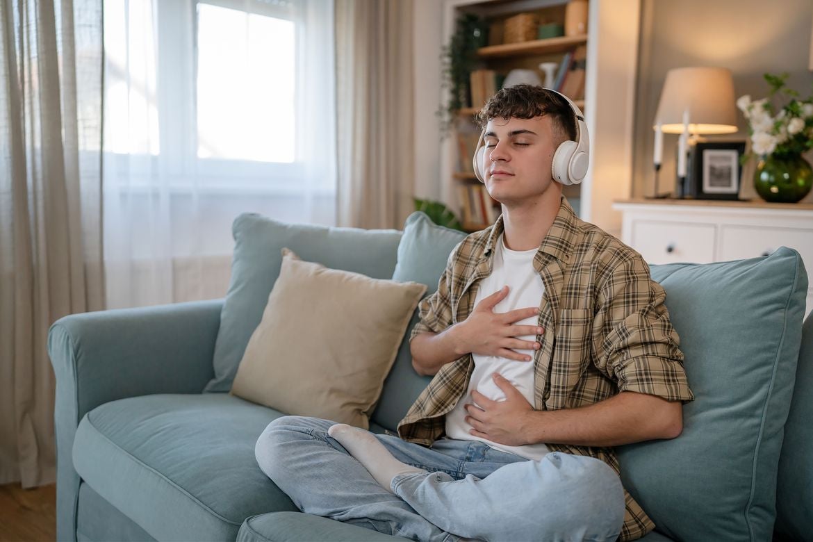 Young man wearing headphones sits cross-legged on a couch, eyes closed, with one hand on his chest and the other on his abdomen.