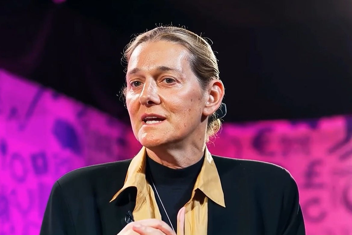 Head-and-shoulders photo of Martine Rothblatt, in black jacket and yellow top, speaking in front of magenta and purple background