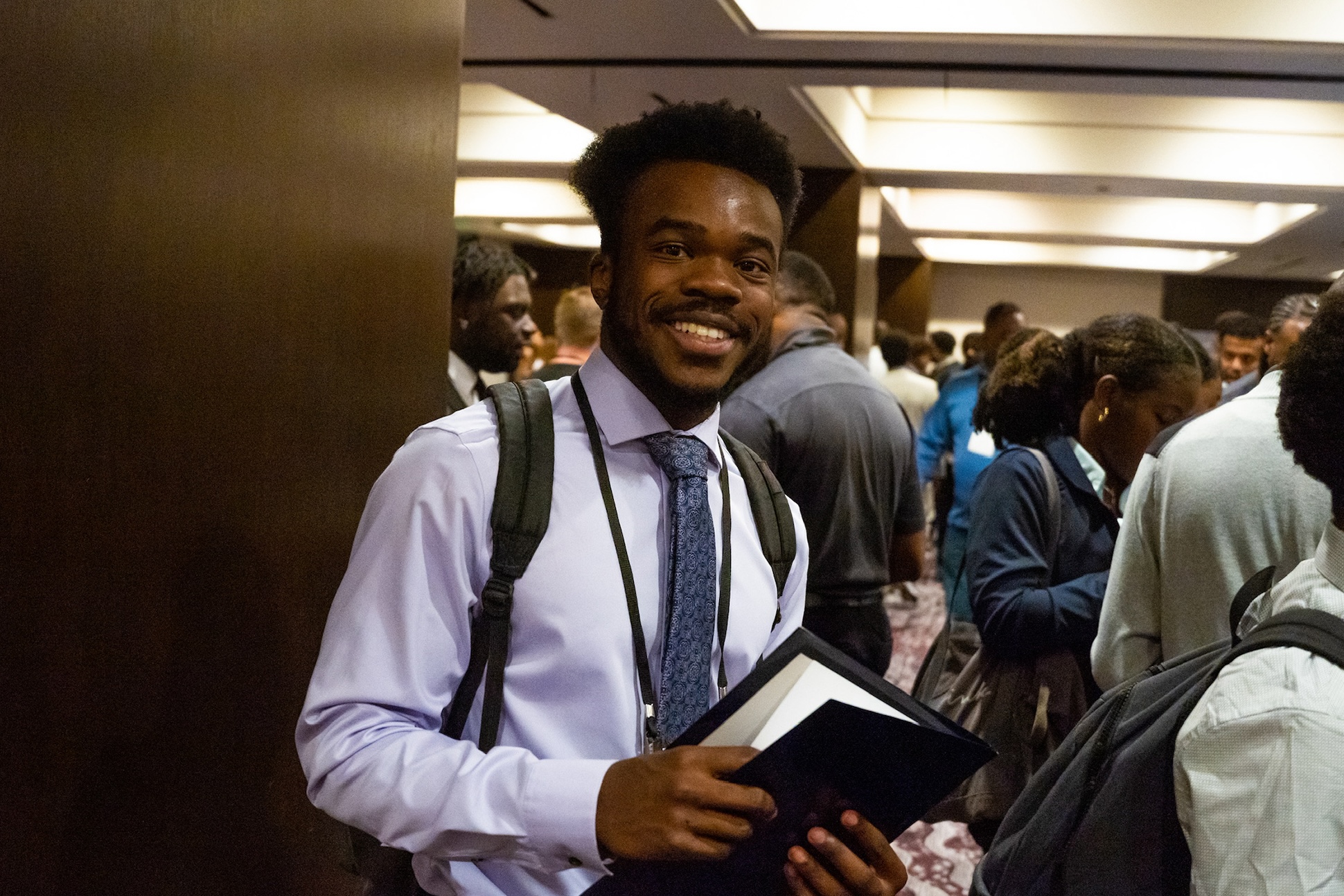 A Black student in a button-up shirt and tie smiles at the camera while holding a folder.