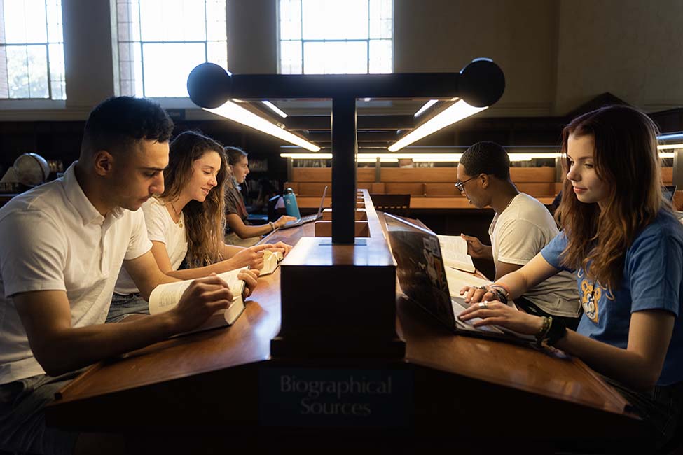 Students study facing each other on opposite sides of lighted library desk, with books and laptops.