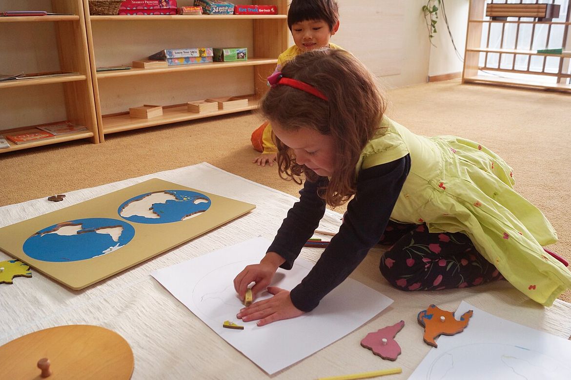 Preschool-aged girl in yellow dress plays on the floor with geography shape puzzle as young boy watches behind her