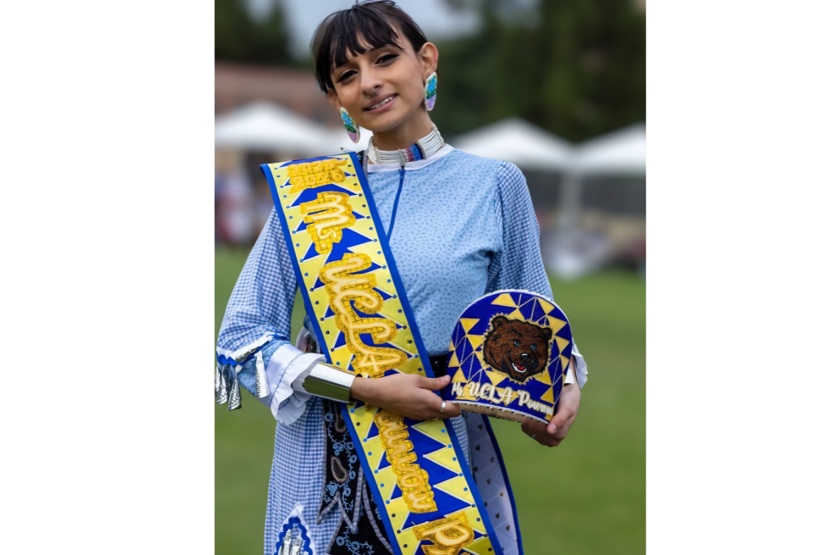 A young woman in a light blue outfit smiles while wearing a “Miss UCLA Powwow” sash and holding a decorated item with a bear design at an outdoor event.