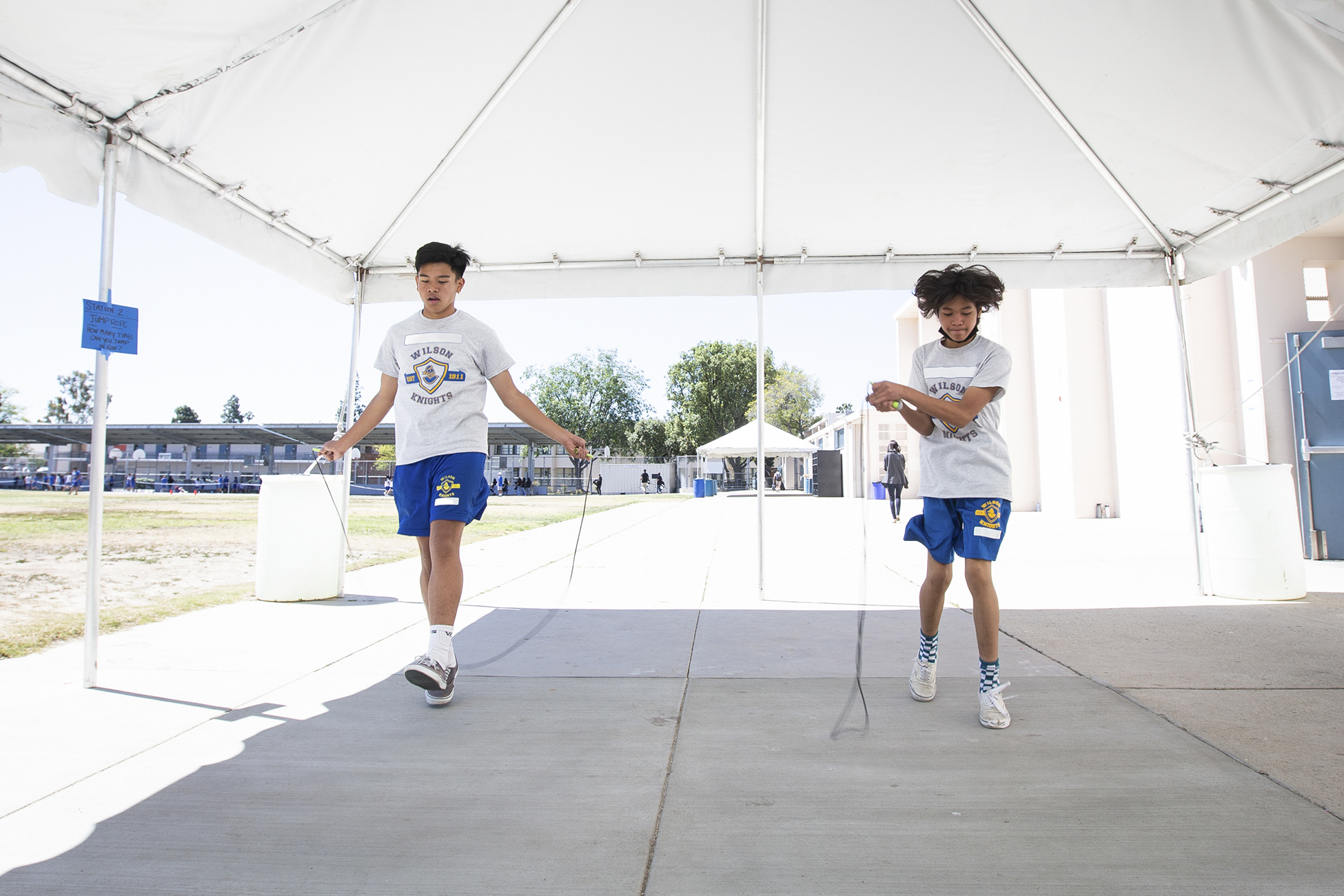 Two teen students, wearing grey and white PE uniforms with a school logo, jump rope under a canopy