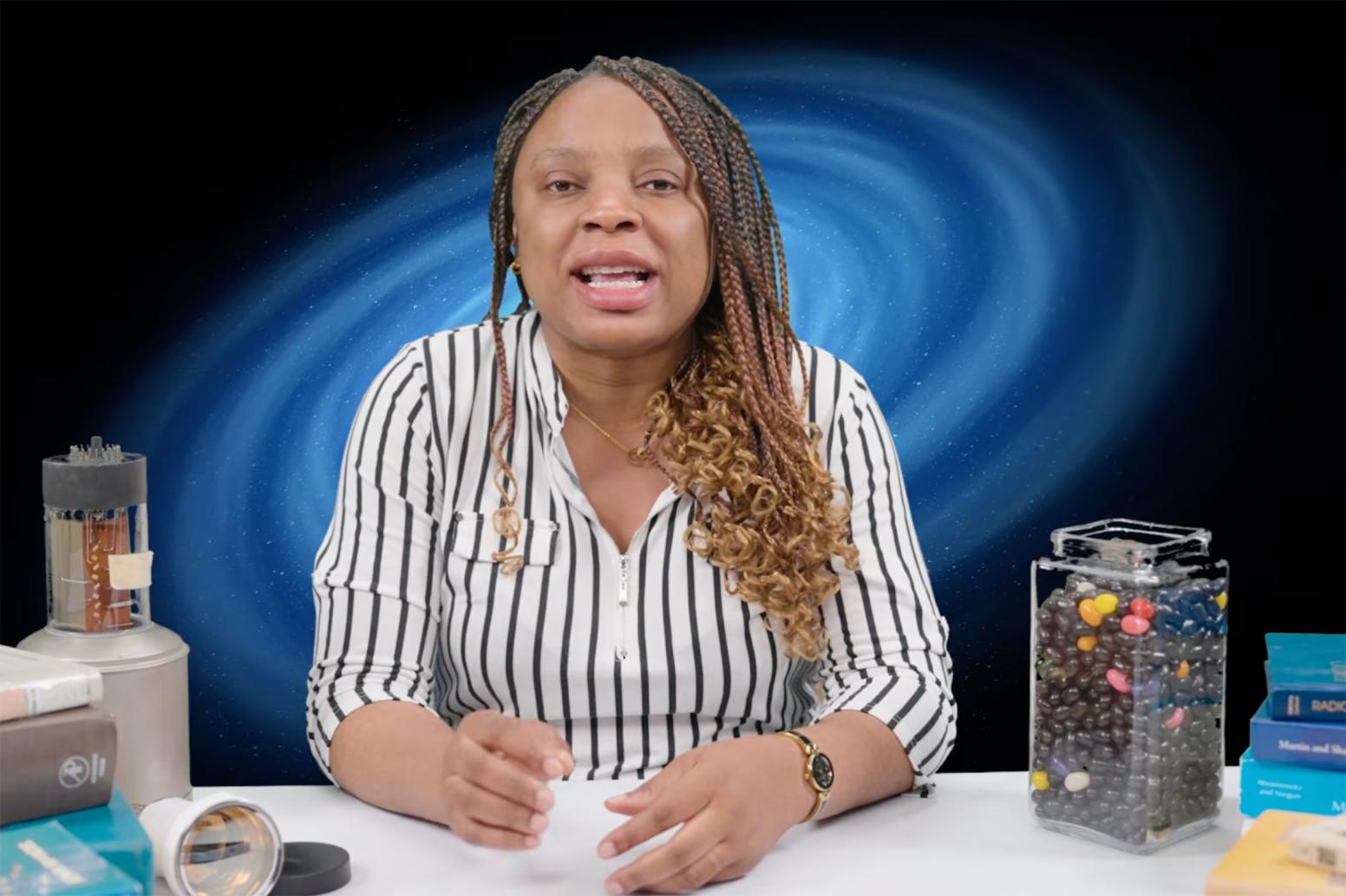 Alvine Kamaha, in a striped blouse sits at a table, speaking toward the camera with a blue abstract backdrop behind her.