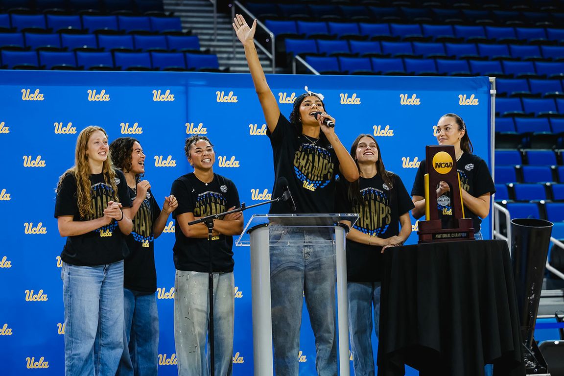Six women in black T-shirts and jeans stand in front of a UCLA step-and-repeat banner