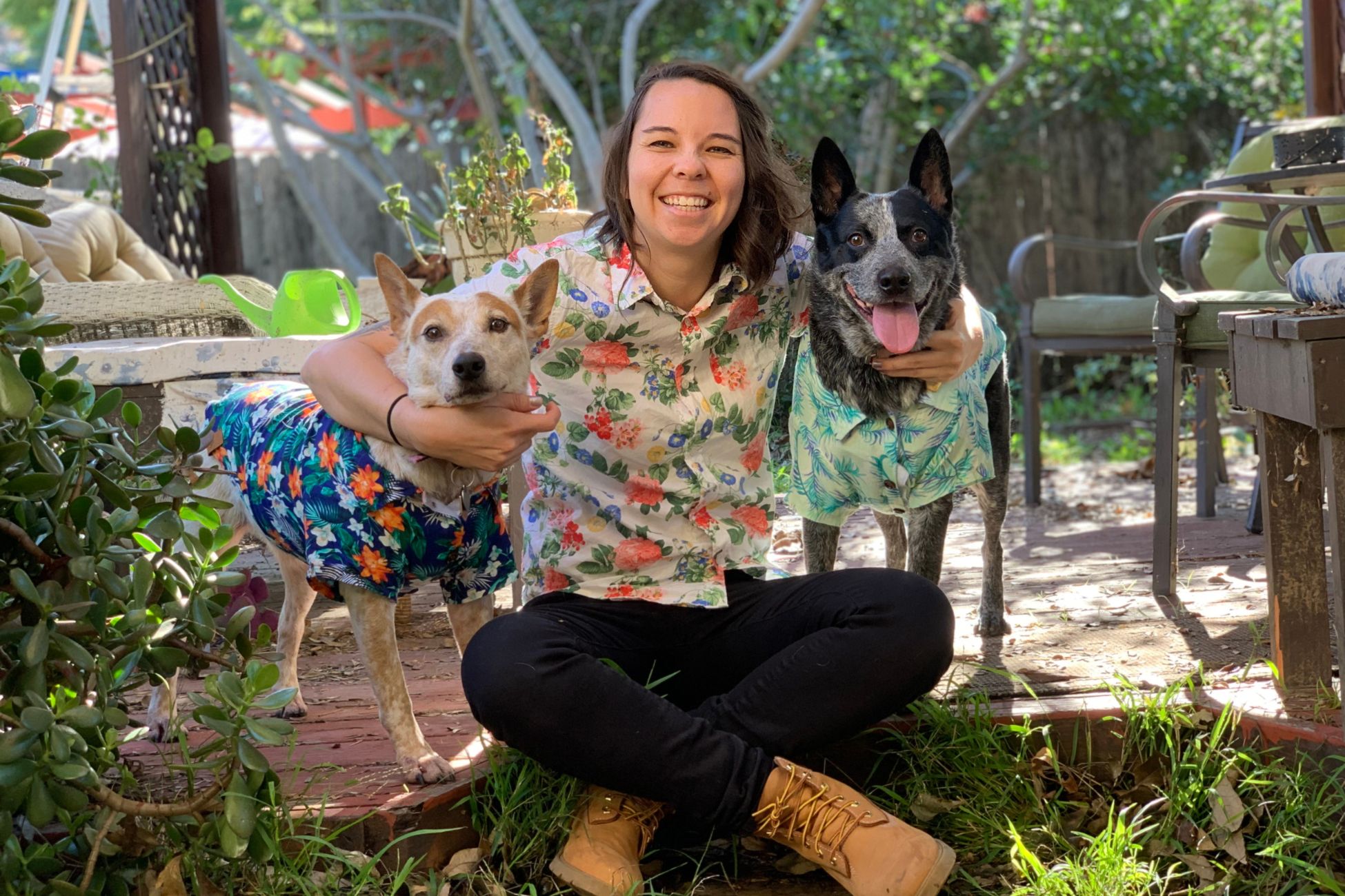 A young woman posing with her two dogs, all wearing floral collared shirts outdoors. 