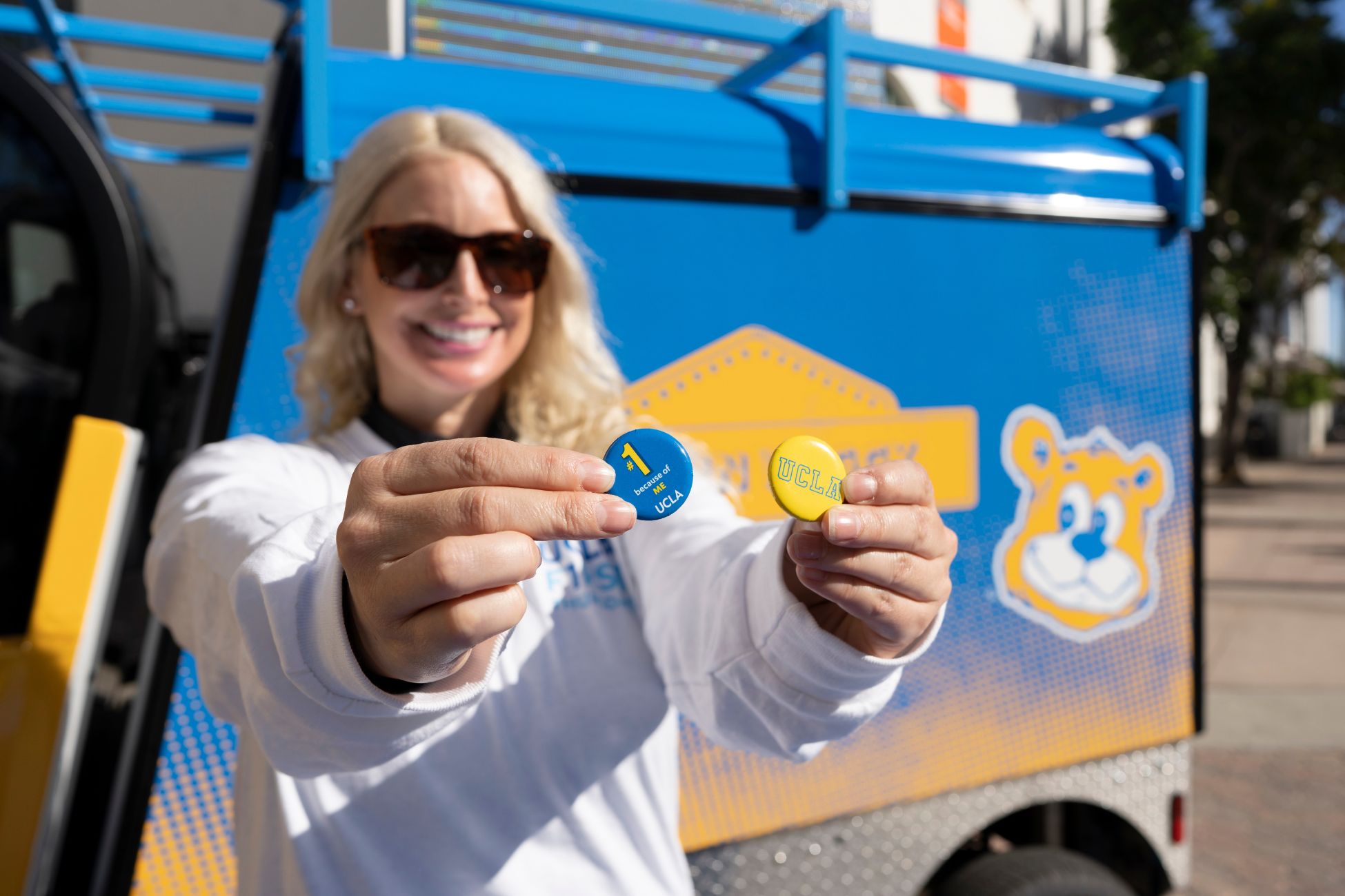 Blonde woman wears sunglasses, holds out 2 UCLA spirit buttons and stands in front of Bruin Buggy