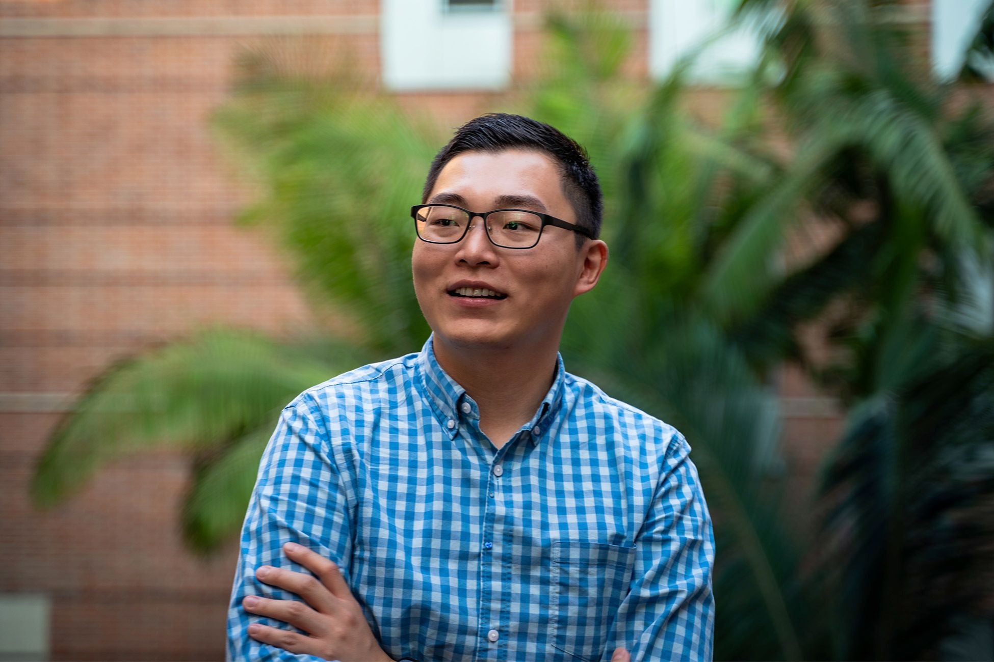 A smiling person stands outdoors in front of blurred greenery and a brick building. Their arms are casually crossed as they look off-camera.