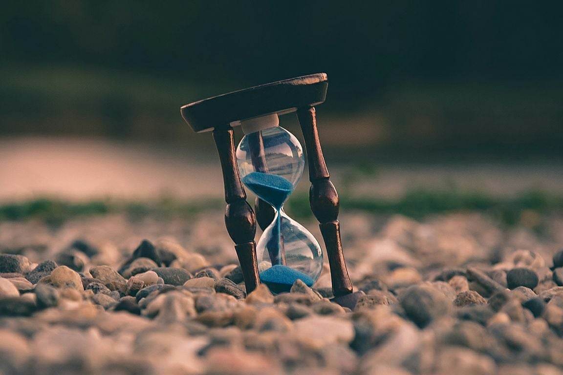 A wooden hourglass with blue sand slowly falling through its glass chamber sits among small rocks and pebbles in an outdoor setting