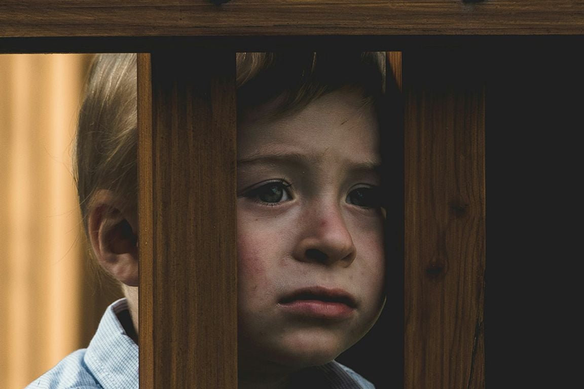 A young child with blonde hair peers through the vertical slats of a wooden railing, their expression somber and tearful.