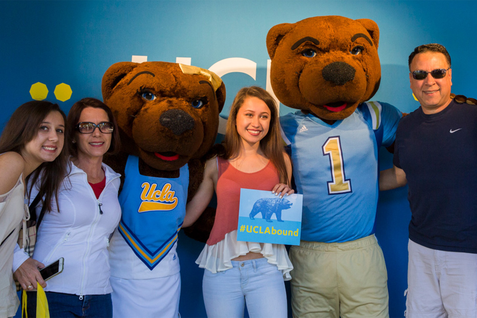 Student holding a #UCLAbound poster next to Joe and Josie Bruin with her family at Bruin Day