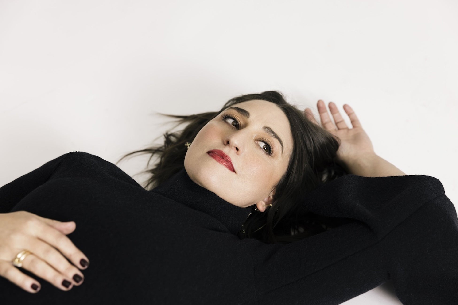 Woman with dark hair and red lipstick lies on a white surface, wearing a black turtleneck sweater and gold rings, looking to the side.