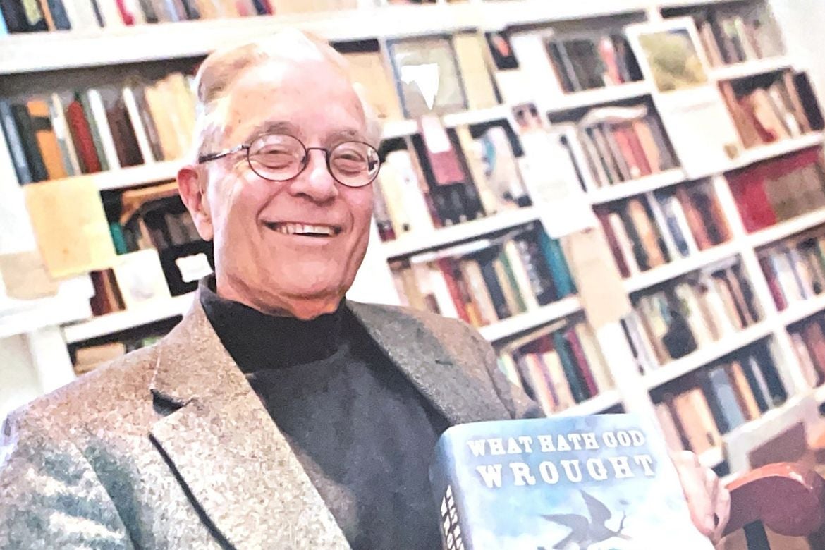 Daniel Walker Howe, in gray jacket and black shirt, smiles as he holds "What Hath God Wrought" book