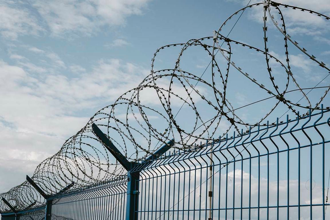 Coils of razor wire atop a blue metal security fence against a partly cloudy sky.