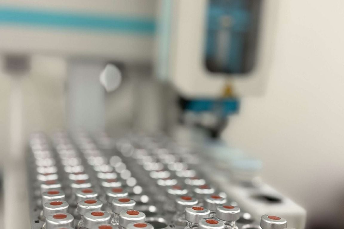 Rows of silver-capped laboratory vials are arranged in a tray, with automated laboratory equipment visible in the background.