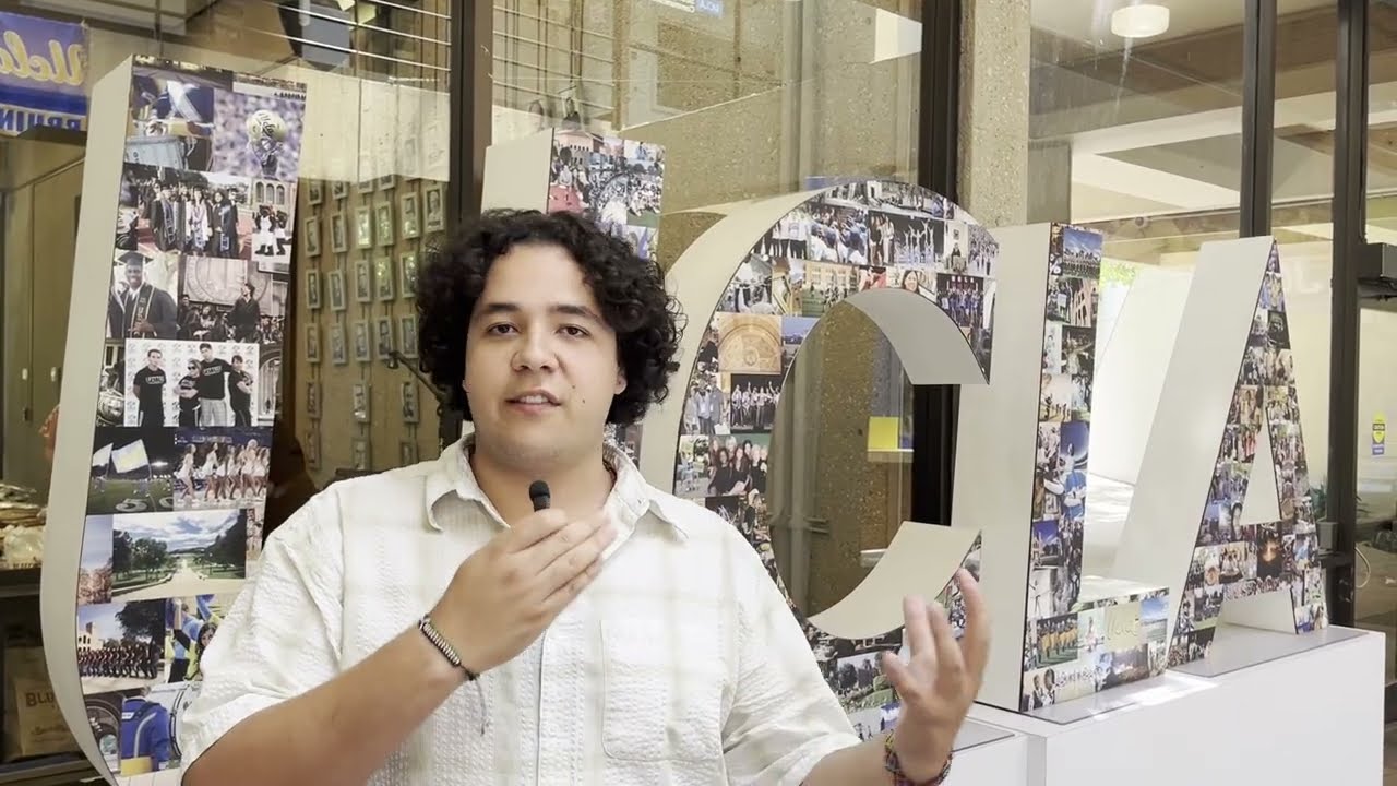 A person holds a microphone in front of a large collage-covered letter display inside a building lobby.