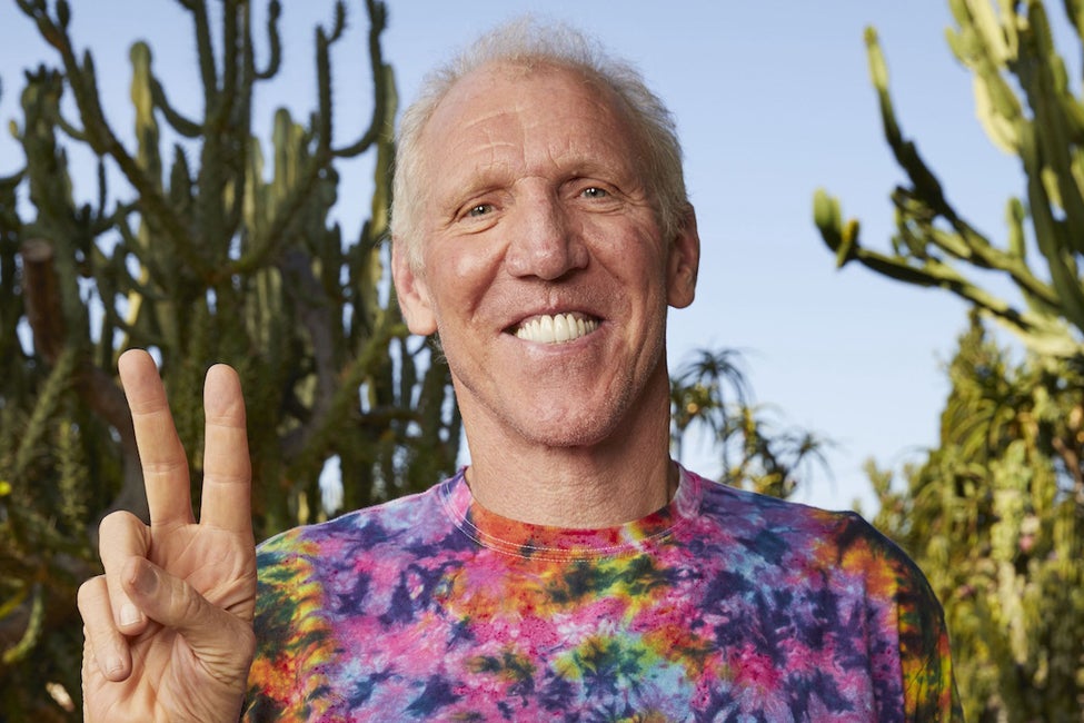 A photo of Bill Walton, wearing a tie dye shirt holding up a peace sign in a cactus garden.