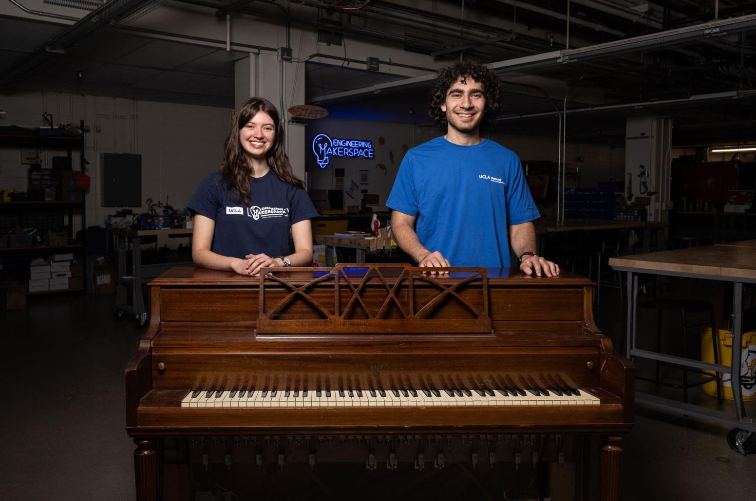 Karina Bender, left, and George Grigoryan wear blue T-shirts as they stand behind a wooden player piano in an engineering makerspace, smiling, with workbenches, tools and a glowing sign behind them.