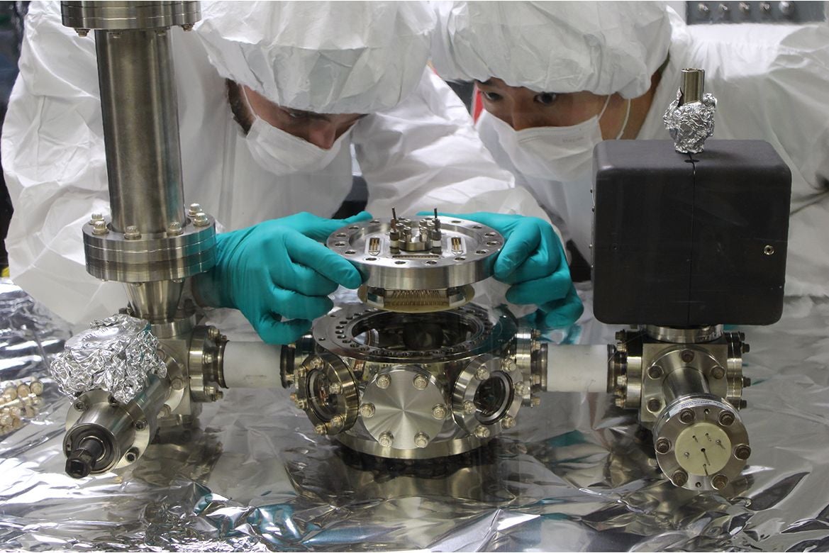 Michael Bareian, left, and Yiyang Zhi in cleanroom suits and gloves carefully install a circular scientific instrument onto a stainless-steel vacuum chamber surrounded by precision laboratory equipment.