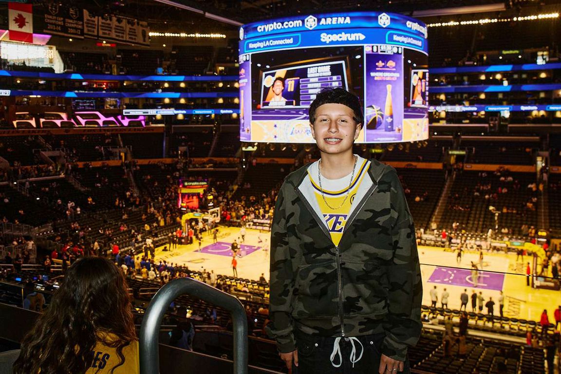 Young basketball player Ava Hernandez smiles courtside at a Lakers game at Crypto.com Arena, with the court and scoreboard visible below.