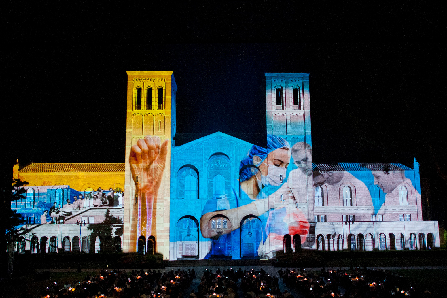People sit in front of Royce Hall at night and watch as the facade is lit by a light show that depicts health and medical professionals.