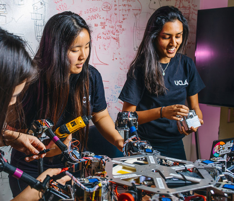 Three female engineering students use power tools to work on a robot.