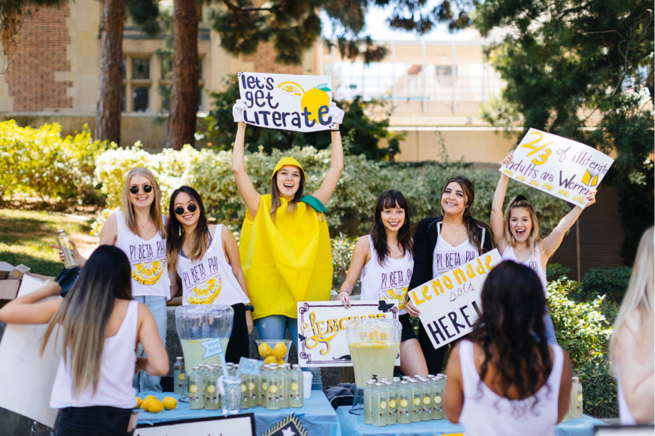 A group of female students are selling lemonade as a fundraiser and holding signs that say “let’s get literate.”