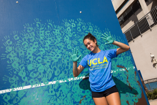 A female student with paint on her hands stands in front of a wall that is covered by her hand prints.