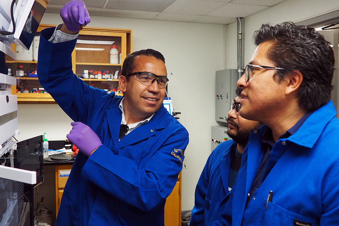 Professor Osvaldo Gutierrez in blue lab coats and safety googles looks on with two fellow lab technictians.