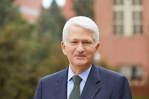 Chancellor Gene Block stands in front of a blurry background composed of red brick and green trees.