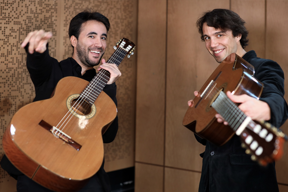 Two men in black shirts hold guitars pointed toward camera and smile; one points hand at camera