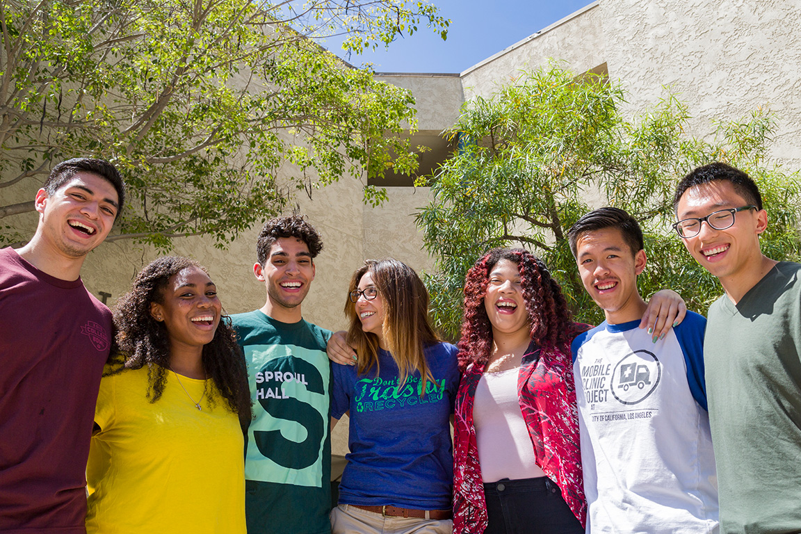 Seven students stand in a group in front of trees and a building as they smile at the camera.